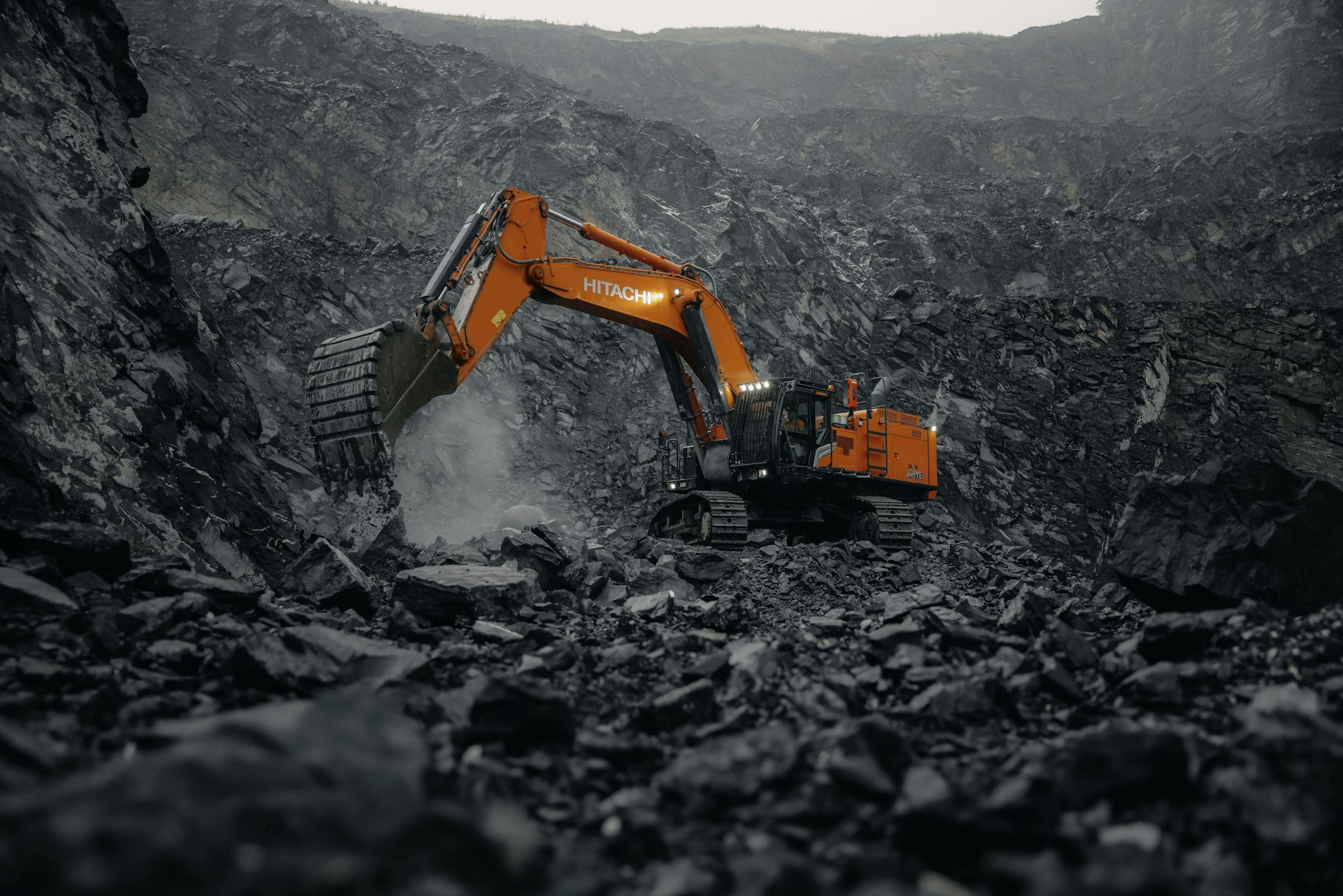 An orange hydraulic excavator digging into a dark, rocky pit wall, with dust rising from the bucket.